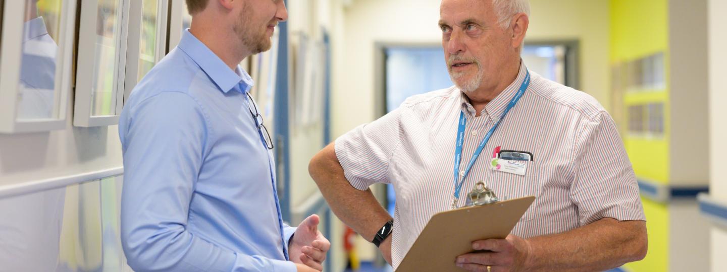 Two men talking over a clipboard in a hospital corridor